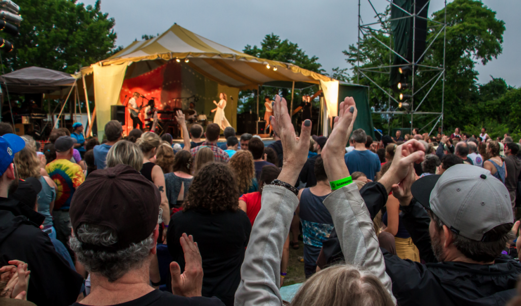 An image of a the crowd in front of a stage at the Great Hudson River Revival music festival.  People in the crowd are raising their arms above their heads to clap.  On stage, the band Lake Street Dive is performing.