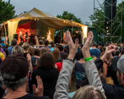 An image of a the crowd in front of a stage at the Great Hudson River Revival music festival. People in the crowd are raising their arms above their heads to clap. On stage, the band Lake Street Dive is performing.