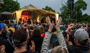An image of a the crowd in front of a stage at the Great Hudson River Revival music festival. People in the crowd are raising their arms above their heads to clap. On stage, the band Lake Street Dive is performing.