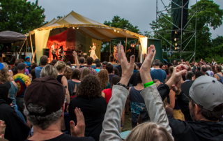 An image of a the crowd in front of a stage at the Great Hudson River Revival music festival. People in the crowd are raising their arms above their heads to clap. On stage, the band Lake Street Dive is performing.