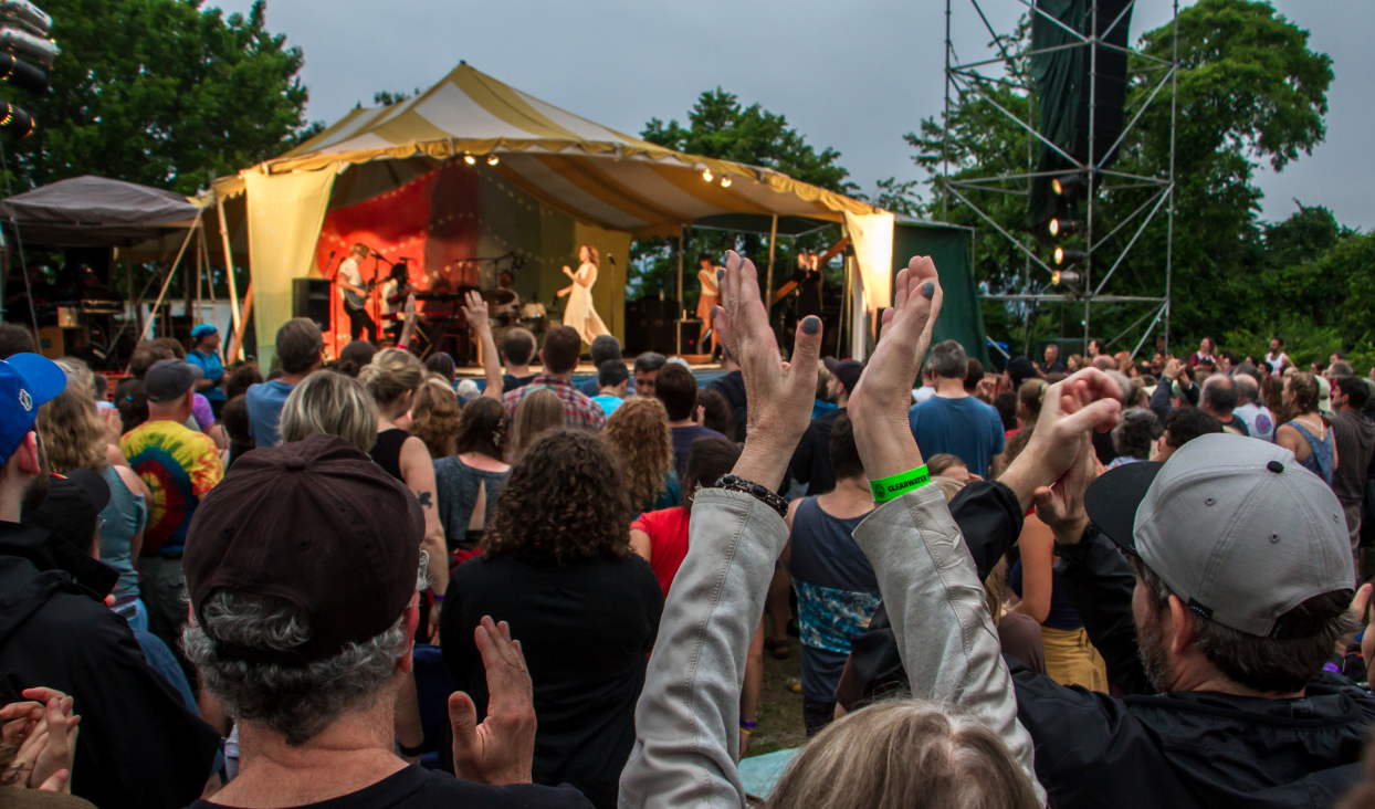 An image of a the crowd in front of a stage at the Great Hudson River Revival music festival. People in the crowd are raising their arms above their heads to clap. On stage, the band Lake Street Dive is performing.