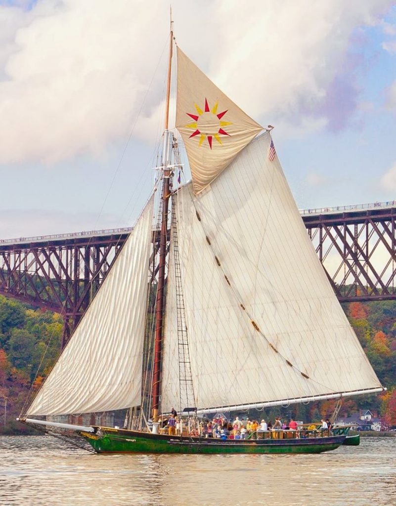 An image of the Hudson River Sloop Clearwater sailing by the Walkway over the Hudson near Poughkeepsie, new York. The sails are set, including the topsail, which has a red and yellow sun pattern on it.