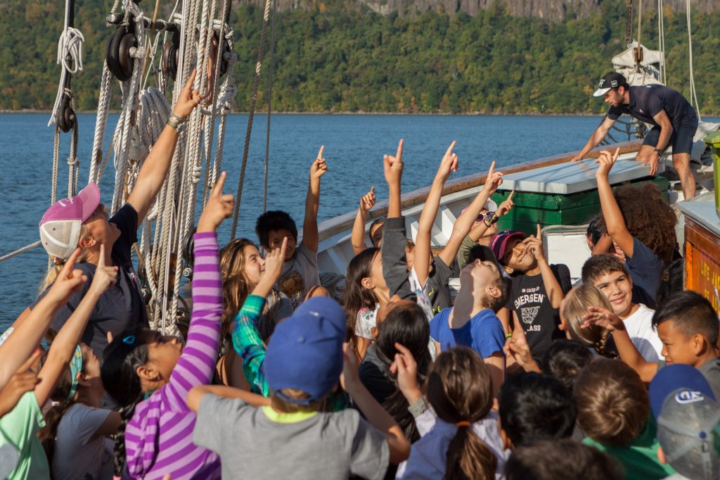 A group of schoolchildren and educators pointing upwards while seated on the port deck of the sailboat Hudson River Sloop Clearwater on a sunny day.